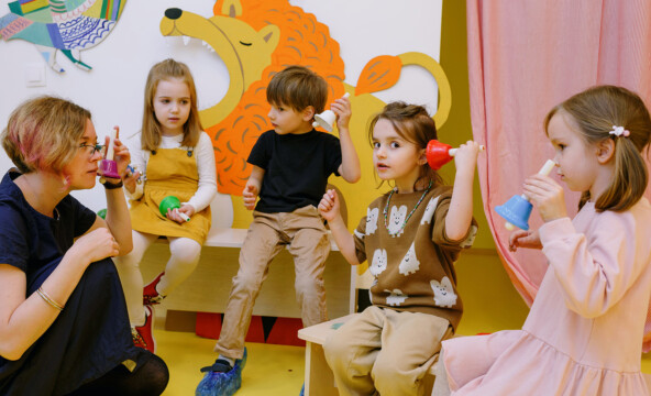 En grupp barn ringer i klockor tillsammans med förskoleläraren. Det finns ett stort gult lejon på klassrumsväggen.