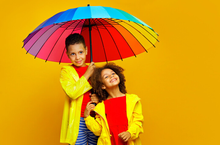 Två glada barn under ett regnbågsfärgad paraply.