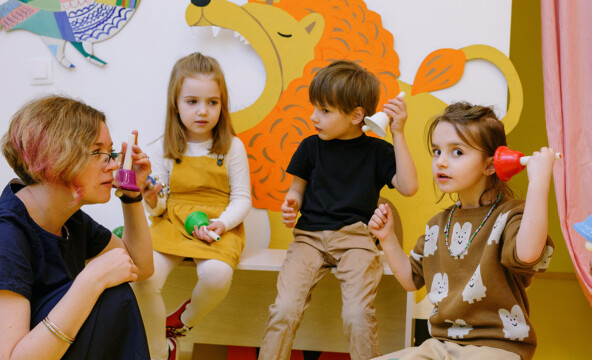 Lapsiryhmä soittaa yhdessä soittokelloja varhaiskasvatuksen opettajan kanssa. Luokan seinällä on iso keltainen leijona.