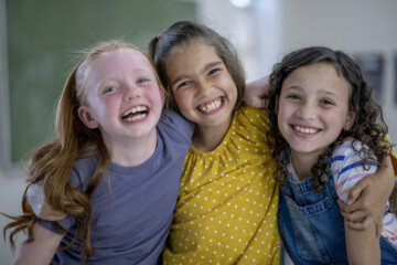 Kolme iloista tyttöä koululuokassa halaavat toisiaan.