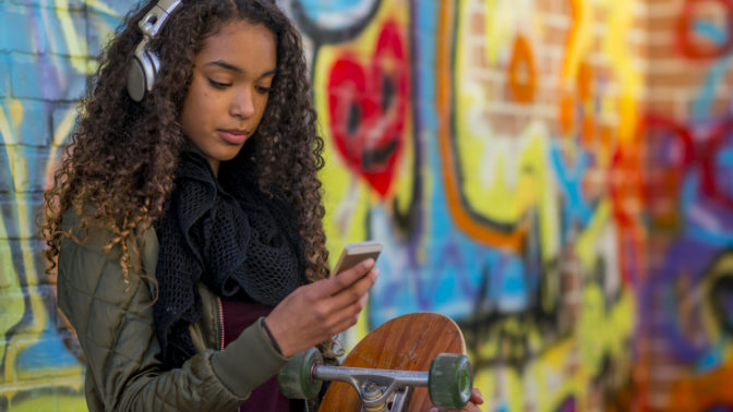 Kiharatukkainen, tumma tyttö näppäilee matkapuhelintaan nojaillen värikkääseen graffitiseinään.