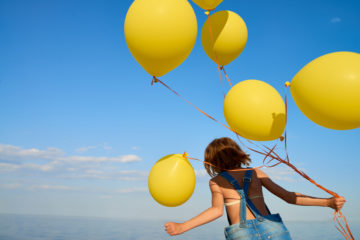 Lapsi selin kädessään keltaisia ilmapalloja, taivas taustalla