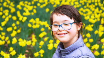 Tyttö, jolla on downin syndrooma, hymyilee kukkapellon edessä.