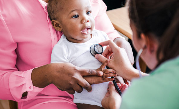 På bilden sitter ett barn i sin förälders famn medan hälsovårdaren lyssnar på barnets bröstkorg med stetoskop.