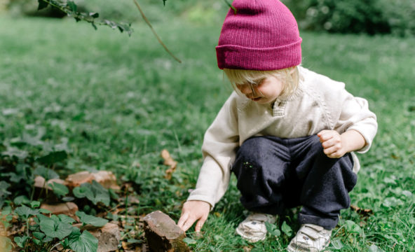 A child with a pink beanie is playing with a stone on a grass yard.