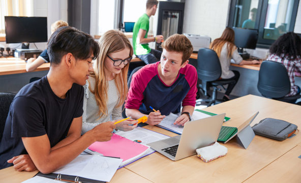 Three young people doing a group assignment in a computer class, with other young people in the background.
