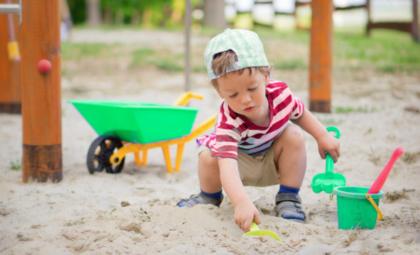 A child plays in a sandbox by a green children’s plastic wheelbarrow, with some lawn in the background.