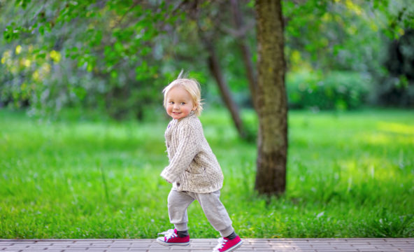 A child dressed in light colours runs along a walkway, with green grass and a tree in the background.