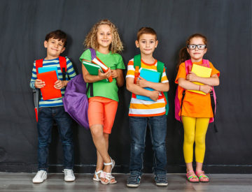 Lapsen oikeuksien viikko 2020: Neljä lasta värikkäissä vaatteissa seisoo liitutaulun edessä sylissään koulukirjoja.