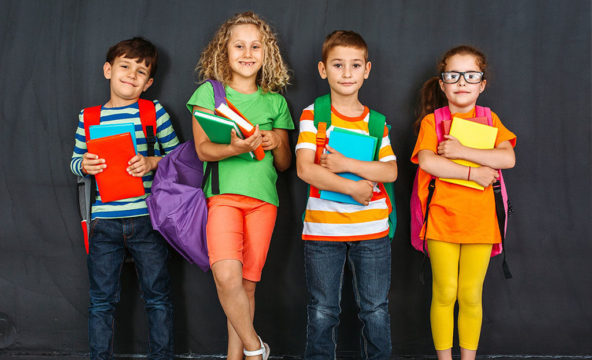 Lapsen oikeuksien viikko 2020: Neljä lasta värikkäissä vaatteissa seisoo liitutaulun edessä sylissään koulukirjoja.