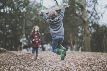 A child hangs his hands on a wooden swing while another is watching in the background.
