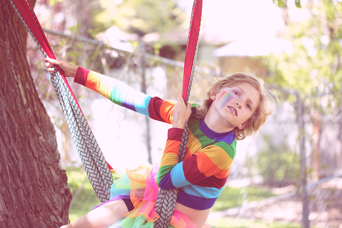 Muunsukupuolinen lapsi leikkimässä ulkona sateenkaaren värisessä paidassa. Poskeen on maalattu pride-lippu
