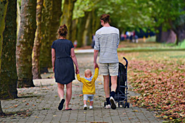 Äiti, pieni taapero ja isä kävelevät kaikki käsi kädessä syksyistä puistotietä. Lapsi on keskellä ja isä työntää lastenvaunuja.