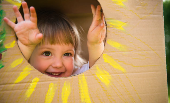 Leikki-ikäinen tyttö kurkistaa pahvilaatikosta, johon on maalattu aurinko.