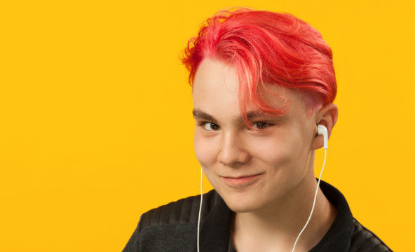 Pinkkitukkainen, teini-ikäinen poika kuuntelee kuulokkeilla hymyillen musiikkia.