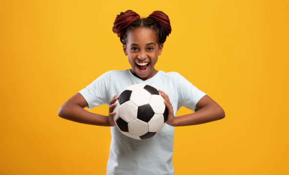 Naurava, tummaihoinen tyttö on heittämässä jalkapalloa kohti kameraa.