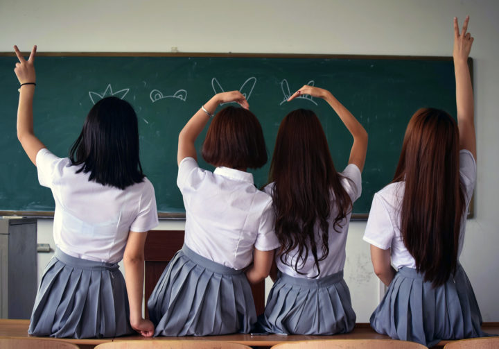 Neljä koulupukuun pukeutunutta tyttöä pelleilee liitutaulun edessä selät kameraan päin.