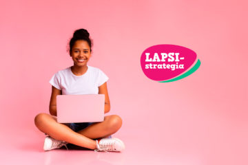 Tummaihoinen, iloinen, nuori tyttö istuu lattialla tietokone sylissään vaaleanpunaista taustaa vasten.